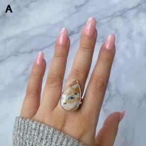 May include: A teardrop-shaped ring with a silver band. The ring features a large, polished stone with a mix of beige, orange, and black patterns. The ring is displayed on a hand with long, pink nails, against a marble background.