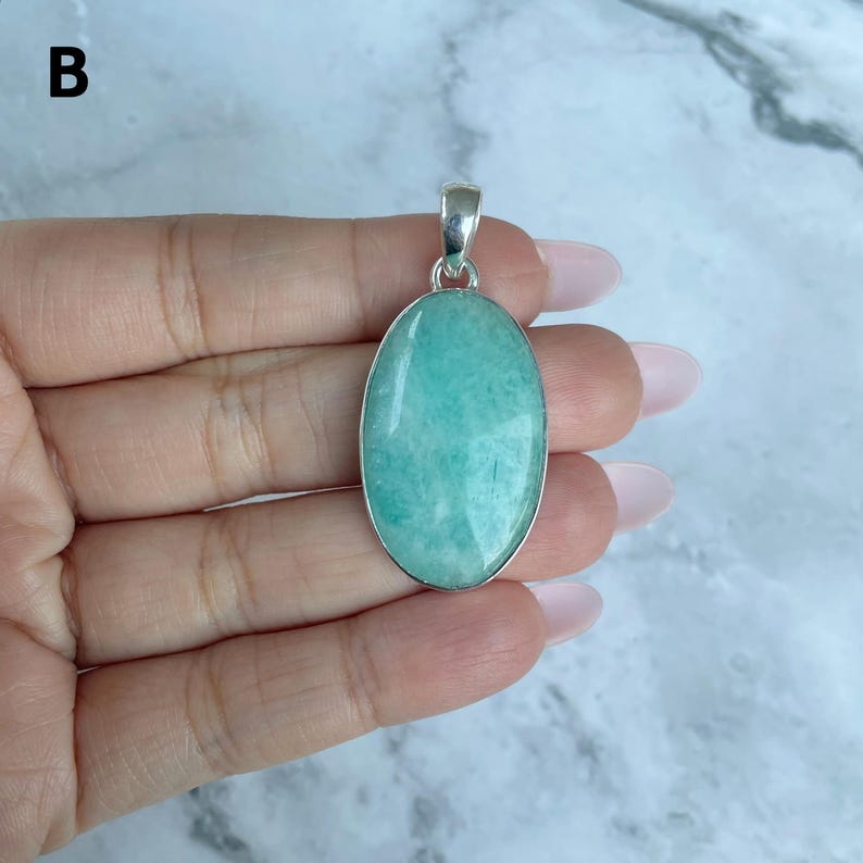 May include: An oval-shaped pendant featuring a smooth, polished light green gemstone set in a silver-toned metal bezel. The pendant includes a small silver-toned bail at the top for attachment to a chain or cord. The letter B is visible in the corner.
