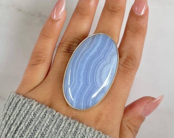 May include: A large, oval-shaped ring featuring a polished, light blue gemstone with a banded pattern. The gemstone is set in a silver-colored metal band. The ring is shown on a hand wearing a gray sweater.