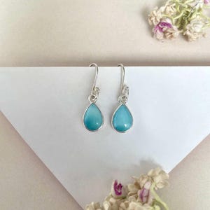 May include: A pair of silver-toned drop earrings with teardrop-shaped, light blue gemstones. The earrings are displayed on a white surface, with delicate floral accents in the background.