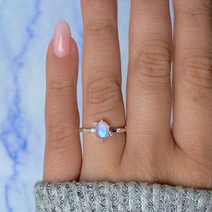 Puede incluir: Un anillo de plata con una piedra lunar ovalada e iridiscente como piedra central. El anillo tiene una banda delicada con pequeñas piedras de acento a cada lado de la piedra lunar. La piedra lunar muestra colores azules y blancos.