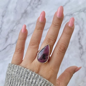 May include: A silver ring featuring a large, teardrop-shaped gemstone. The gemstone is a deep pink color with dark inclusions. The ring is set in a simple silver band. The ring is shown on a hand with pink nail polish.
