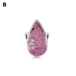 May include: A silver ring featuring a large, teardrop-shaped pink druzy stone. The stone has a textured surface with varying shades of pink and white. The ring's band is a simple, polished silver.