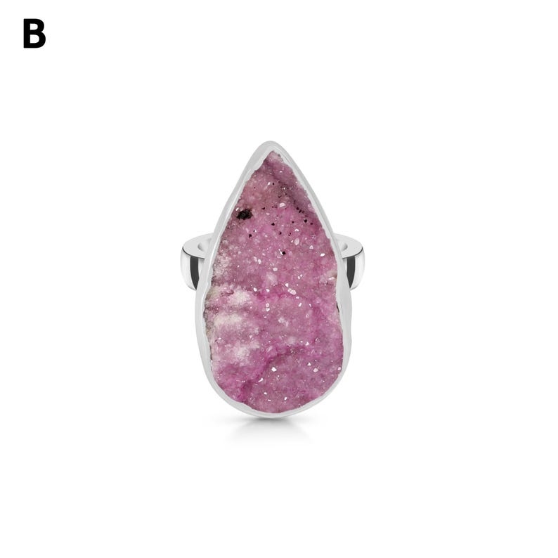 May include: A silver ring featuring a large, teardrop-shaped pink druzy stone. The stone has a textured surface with varying shades of pink and white. The ring's band is a simple, polished silver.