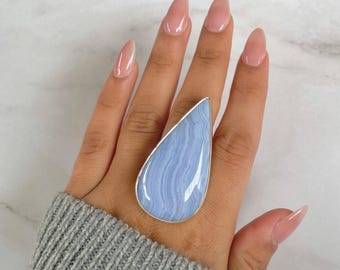 May include: A large, teardrop-shaped ring featuring a pale blue gemstone with a silver-colored bezel. The stone has a striped pattern. The ring is displayed on a hand wearing a gray sweater. The letter "A" is in the upper left corner.