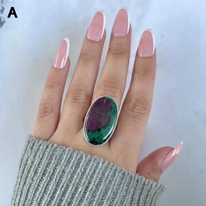 May include: A silver ring featuring a large, oval-shaped cabochon stone with a distinct color split of deep magenta-red and dark forest green. The stone is set in a smooth, polished silver bezel, showcasing a natural, unrefined mineral texture.