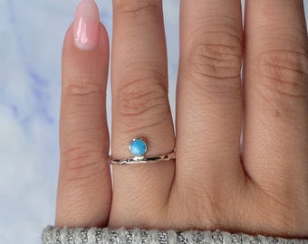 May include: A silver ring with a small, round, light blue stone set on a textured band. The ring is displayed on a finger, showcasing its delicate design and color contrast. The ring's minimalist style makes it suitable for everyday wear.