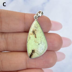 Pu&ograve; includere: Un ciondolo a forma di goccia con una pietra verde lime pallido e inclusioni di matrice marrone scuro, incastonato in una montatura in metallo color argento lucido con una contromaglia. La lettera C &egrave; visibile nell'angolo in alto a sinistra.