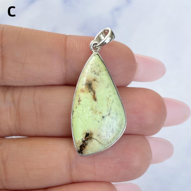 Pu&ograve; includere: Un ciondolo a forma di goccia con una pietra verde lime pallido e inclusioni di matrice marrone scuro, incastonato in una montatura in metallo color argento lucido con una contromaglia. La lettera C &egrave; visibile nell'angolo in alto a sinistra.