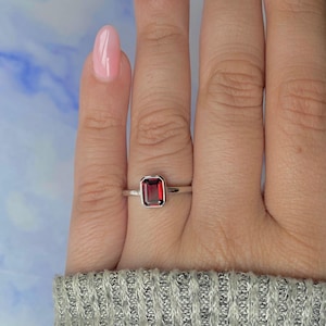 May include: A silver ring with a rectangular, deep red gemstone. The ring is set on a hand with pink nail polish, against a blue and white background. The ring's design is simple and elegant, showcasing the vibrant color of the stone.