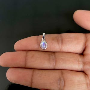 May include: A small, oval moonstone pendant with a silver bail. The moonstone displays a milky, iridescent quality, with flashes of blue and white. The pendant is held in a hand against a dark backdrop.