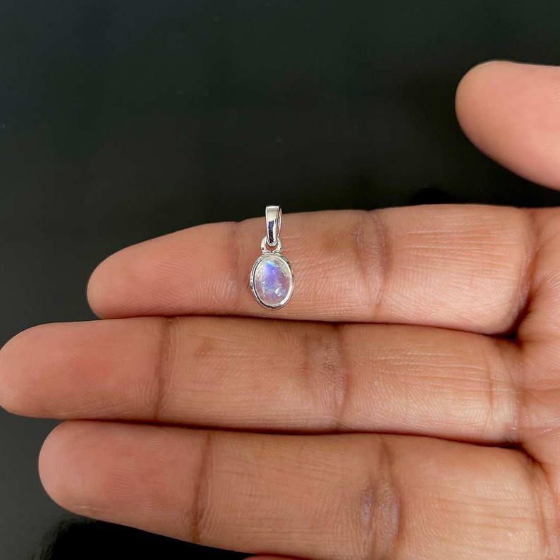 May include: A small, oval moonstone pendant with a silver bail. The moonstone displays a milky, iridescent quality, with flashes of blue and white. The pendant is held in a hand against a dark backdrop.
