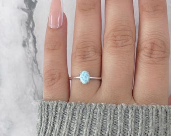 May include: A silver ring featuring an oval, light blue gemstone. The ring is set on a thin band and is displayed on a hand wearing a gray sweater. The gemstone has a marbled appearance.