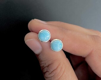May include: A pair of round stud earrings featuring polished, sky-blue Larimar gemstones set in simple silver bezels. Each circular stone displays natural white marbling patterns within the light blue surface, secured by a smooth metallic rim.