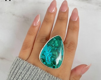 Könnte beinhalten: Ein tropfenförmiger Ring mit einem polierten Malachit-Stein, der wirbelnde Muster in tiefem Waldgrün und leuchtendem Türkis aufweist. Der Edelstein ist in eine glatte, silberfarbene Metallfassung eingefasst und bildet ein auffälliges Schmuckstück am Finger.