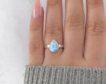 May include: A silver ring featuring a pear-shaped, light blue gemstone. The ring is displayed on a hand, showcasing the polished stone's color and the ring's simple band design. The gemstone has a unique, marbled appearance.