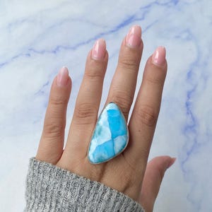 May include: A large, triangular ring featuring a vibrant blue larimar stone with white marbling. The stone is set in a silver-colored metal band. The ring is worn on a hand with manicured nails, and a gray sweater.