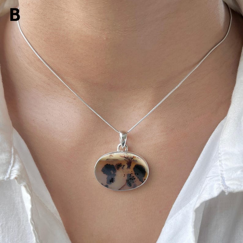 Puede incluir: Un collar de plata con una cadena fina y un colgante ovalado. El colgante contiene una piedra lisa de color claro con inclusiones dendr&iacute;ticas oscuras en forma de &aacute;rbol. La letra B es visible en la esquina superior izquierda de la imagen.