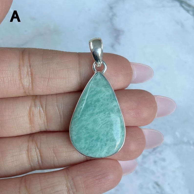 May include: A teardrop-shaped pendant featuring a smooth, polished light green stone with subtle white marbling, encased in a polished silver-toned metal setting with a small bail at the top, marked with the letter A in the upper left corner.