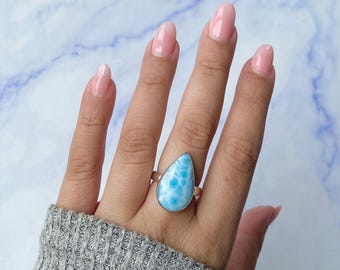 May include: A silver ring featuring a large, teardrop-shaped, light blue stone with white marbled patterns. The stone is set in a smooth metal bezel, and the band is a simple, polished silver wire. The ring is worn on a finger, showcasing the stone.