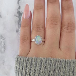 May include: A silver ring with an oval opal gemstone. The opal displays a play of colors, including white, blue, and green. The ring is set on a finger, with a gray sweater in the background.