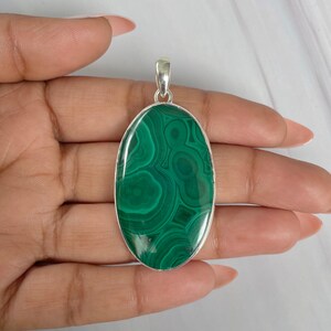 Peut inclure: Pendentif ovale en malachite avec une b&eacute;li&egrave;re argent&eacute;e. La pierre verte polie pr&eacute;sente un motif tourbillonnant de teintes vertes plus claires et plus fonc&eacute;es. Le pendentif est tenu dans une main, mettant en valeur sa taille et son design.