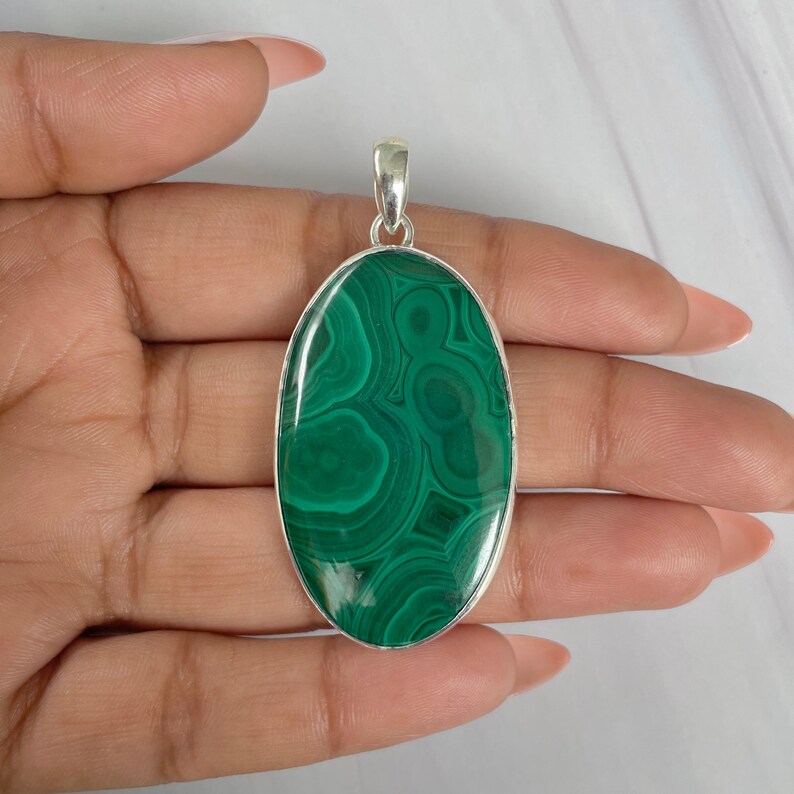 Peut inclure: Pendentif ovale en malachite avec une b&eacute;li&egrave;re argent&eacute;e. La pierre verte polie pr&eacute;sente un motif tourbillonnant de teintes vertes plus claires et plus fonc&eacute;es. Le pendentif est tenu dans une main, mettant en valeur sa taille et son design.