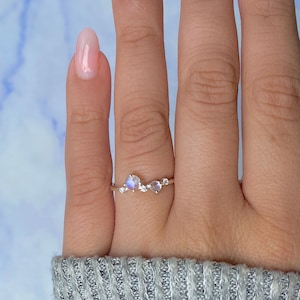 May include: A silver ring featuring a cluster of three gemstones. The central stone is a pearlescent, iridescent moonstone, flanked by two smaller, heart-shaped stones and several small, clear accent stones. The ring is set on a finger.