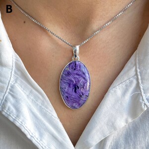 Op de afbeelding: Een ovaalvormige charoiet hanger gezet in een gepolijst zilveren montuur, hangend aan een fijne zilveren ketting. De edelsteen heeft een wervelend, gemarmerd patroon van diepe violette en paarse tinten met subtiele zwarte en witte insluitingen.
