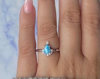 May include: A silver ring featuring a pear-shaped, light blue gemstone, surrounded by a halo of small, clear stones. A single, small clear stone sits above the main stone. The ring is on a finger, with a light blue and white background.