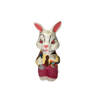 May include: A white rabbit figurine with pink ears and red eyes, wearing a yellow shirt, a brown tie, and pink pants. The rabbit holds a pocket watch and a patchwork jacket. The figurine is a whimsical decorative item.