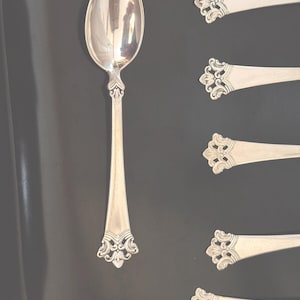 May include: Five silver spoons with ornate handles. The spoons are arranged in a row on a dark surface. The spoon on the left is positioned vertically, while the other four spoons are positioned horizontally.