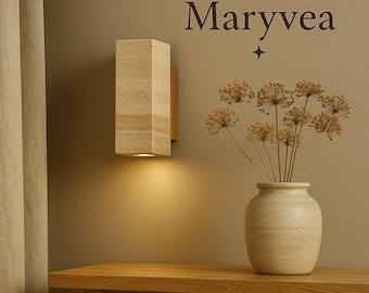 May include: A rectangular, beige stone wall sconce with a warm light. The sconce is mounted on a wooden backing. A vase with dried flowers sits on a wooden table. The word "Maryvea" is above the sconce.