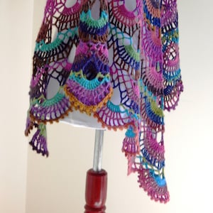 Puede incluir: Un chal de ganchillo hecho a mano y colorido en tonos morado, rosa, azul, verde y marrón. El diseño calado presenta un patrón repetitivo en forma de abanico con bordes festoneados. El chal se exhibe en un maniquí.