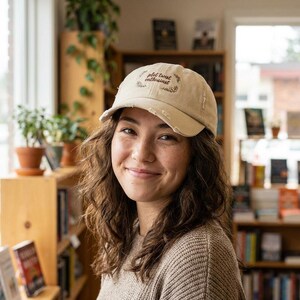 May include: A beige baseball cap with the words "plot twist enthusiast" embroidered on the front. The cap has a distressed appearance. The person wearing the cap is in a bookstore, with bookshelves and plants visible in the background.
