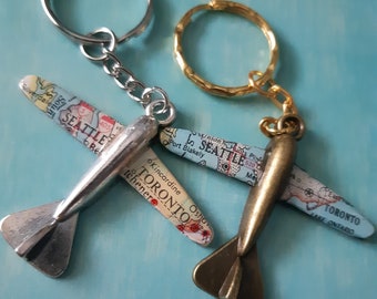 May include: Two silver and bronze airplane keychains with map details. The silver keychain has the text "SEATTLE" and "TORONTO" on it. The bronze keychain has the text "SEATTLE" and "TORONTO" on it.