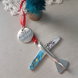 May include: A silver airplane ornament with map-covered wings and a red ribbon. The ornament has the text "no matter where" on a silver disc. A small, teal bottle brush tree is in the background.