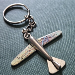 May include: A silver-toned metal airplane keychain featuring wings decorated with a printed map design. The wings display geographical text including the names Moscow and Ottawa. The keychain includes a circular metal split ring attached by a chain.