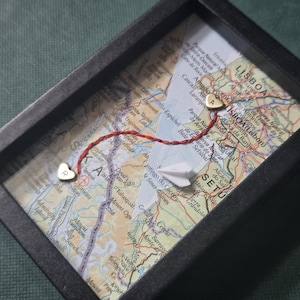 May include: A black rectangular shadow box frame containing a map of Lisbon, Portugal. Two small gold-colored heart charms, one engraved with the letter R and the other with A, are connected by a red thread. A small white paper airplane is included.