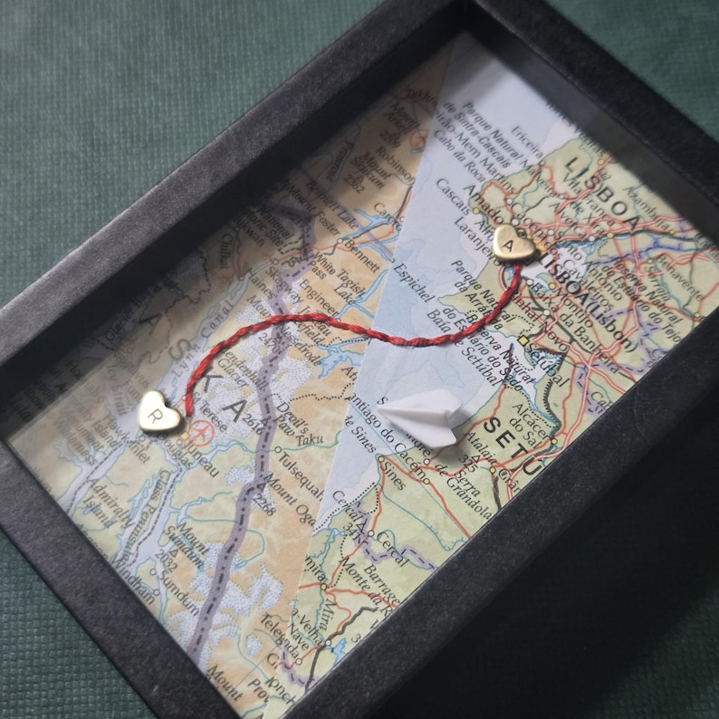 May include: A black rectangular shadow box frame containing a map of Lisbon, Portugal. Two small gold-colored heart charms, one engraved with the letter R and the other with A, are connected by a red thread. A small white paper airplane is included.