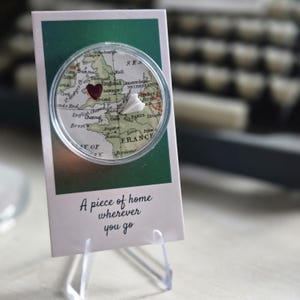 May include: A clear acrylic display stand holding a card with a vintage map design. A heart-shaped charm marks a location on the map. The card reads "A piece of home wherever you go."