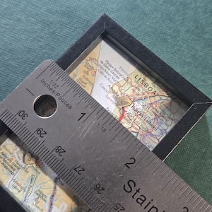May include: A black-framed square display containing a map of Lisbon, Portugal, featuring two small gold heart-shaped charms marked with the letters A and R. A stainless steel ruler is placed over the display, showing a measurement of 2 inches.