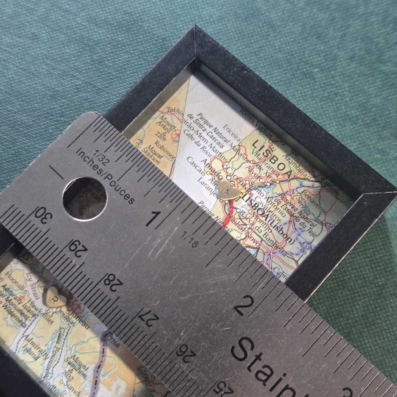 May include: A black-framed square display containing a map of Lisbon, Portugal, featuring two small gold heart-shaped charms marked with the letters A and R. A stainless steel ruler is placed over the display, showing a measurement of 2 inches.