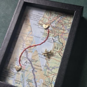 May include: A framed map display featuring two gold-toned heart charms, one marked with the letter A and the other with the letter R, connected by a red thread. A small gold-toned airplane charm is positioned on the map surface near the letter R.