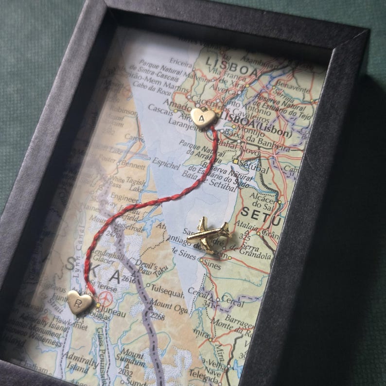 May include: A framed map display featuring two gold-toned heart charms, one marked with the letter A and the other with the letter R, connected by a red thread. A small gold-toned airplane charm is positioned on the map surface near the letter R.