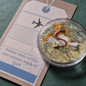 May include: A clear, round keepsake with a map design, a red string, and two heart-shaped charms. A paper airplane and a tag with the text "Distance means so little when someone means so much" are also present.