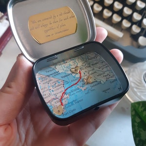 May include: A small, round tin box with a map of Italy inside. The map has a red line drawn on it, connecting two gold hearts. The tin box has a gold inscription on the lid that reads: "We are connected by a red thread and will always be there for each other regardless of place, time or circumstances."