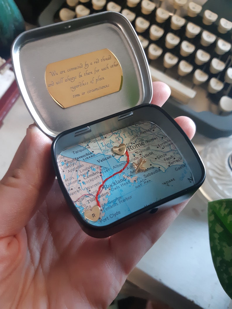 May include: A small, round tin box with a map of Italy inside. The map has a red line drawn on it, connecting two gold hearts. The tin box has a gold inscription on the lid that reads: "We are connected by a red thread and will always be there for each other regardless of place, time or circumstances."