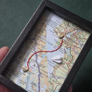 May include: A black shadow box frame containing a map of Alaska and Lisbon, Portugal. A red thread connects two small gold heart charms, one stamped with the letter R and the other with A. A small white paper airplane is placed on the map surface.