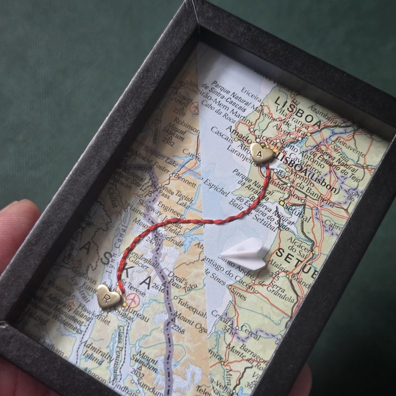 May include: A black shadow box frame containing a map of Alaska and Lisbon, Portugal. A red thread connects two small gold heart charms, one stamped with the letter R and the other with A. A small white paper airplane is placed on the map surface.